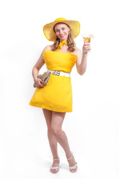 La mujer en vestido amarillo de la almohada, el sombrero con el monedero y la copa de cóctel sonríen en el fondo blanco. Concepto de verano Desafío de almohadas debido al aislamiento en el hogar.