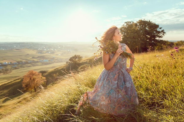 Una mujer con un vestido azul se para en un campo con un ramo de flores en la mano.