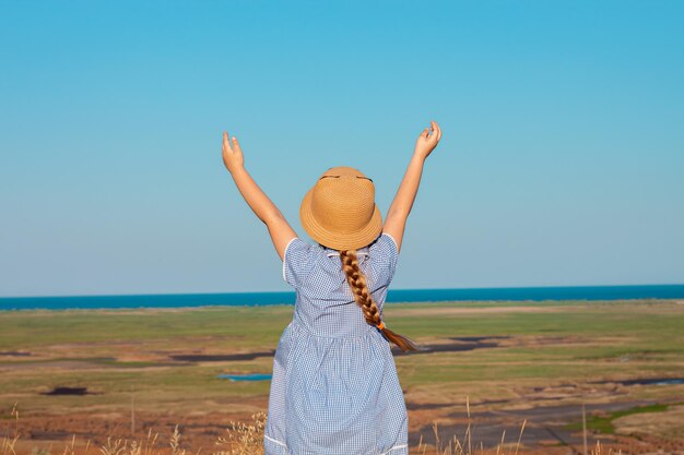 Una mujer con un vestido azul y un sombrero de paja se para en un campo con los brazos levantados en el aire.