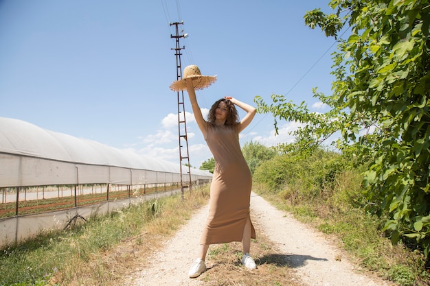 mujer en un vestido beige y sombrero de paja posando en una carretera rural