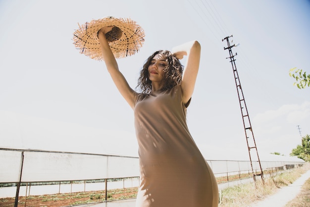 mujer en un vestido beige y sombrero de paja posando en una carretera rural