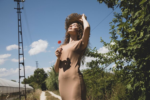 mujer en un vestido beige y sombrero de paja posando en una carretera rural