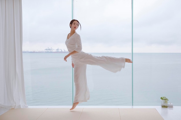 Una mujer con un vestido blanco baila frente a una ventana con vista al océano al fondo.