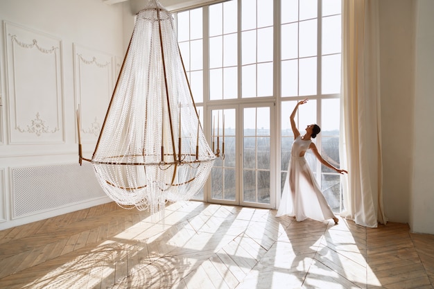 Mujer en vestido blanco baila junto a la ventana en estudio de luz