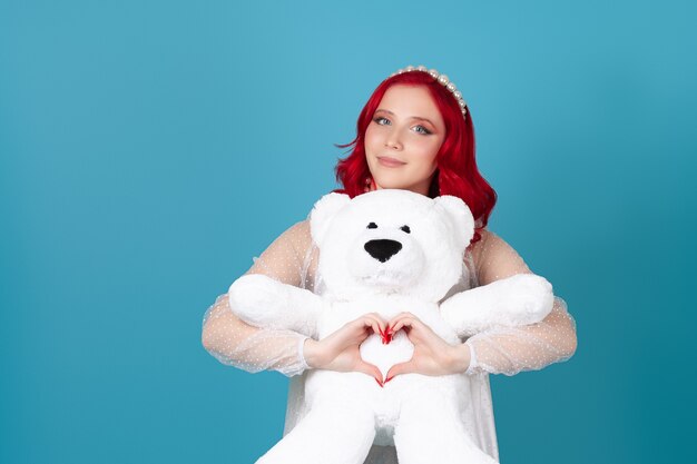 Foto mujer con un vestido blanco con cabello rojo abraza suavemente a un oso de peluche blanco y hace un símbolo de corazón con sus dedos