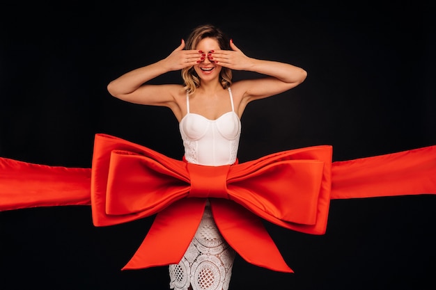 Una mujer con un vestido blanco como regalo se cubre los ojos con las manos sobre una superficie negra envuelta en una cinta festiva