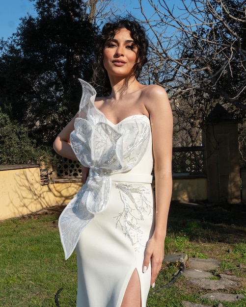 Una mujer con un vestido blanco se para frente a un árbol.