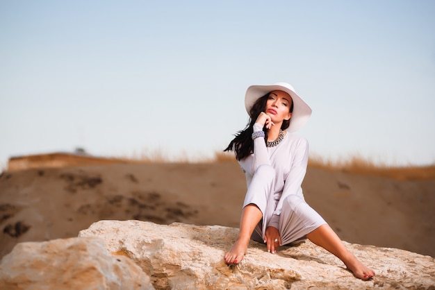 Mujer con vestido blanco sentado en una roca en la playa y con sombrero blanco.