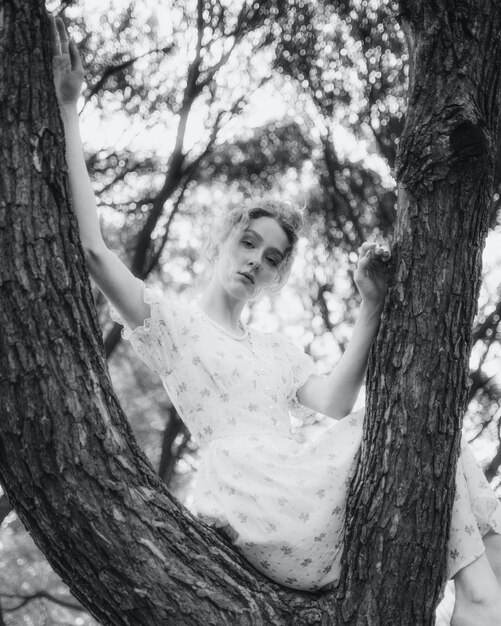 Una mujer con un vestido blanco se sienta en la rama de un árbol y mira a la cámara.