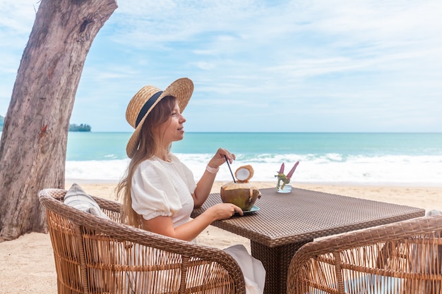 Mujer con vestido blanco y sombrero de paja sentado en la mesa en el café de la playa al aire libre