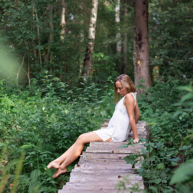 Mujer con vestido corto blanco en el bosque de verano se sienta descalza en un puente de madera