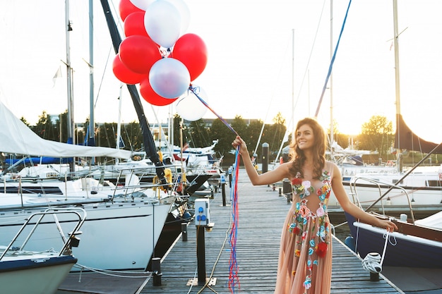 Mujer en vestido hermoso con muchos globos de colores en el muelle del yate