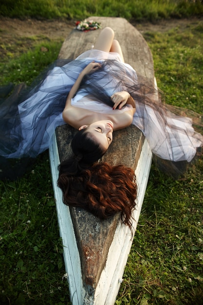 Una mujer con un vestido largo cerca del lago se sienta en un viejo bote, una hermosa mujer bajo los rayos del sol de la tarde en el otoño en el pueblo. Vida rural y moda