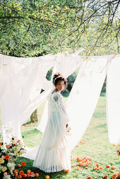 Una mujer con un vestido largo se encuentra cerca de las cortinas blancas en la hierba verde del jardín