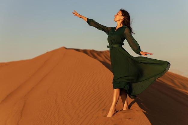 Mujer en vestido largo verde se encuentra en dunas de arena del desierto fondo amarillo naranja puesta de sol