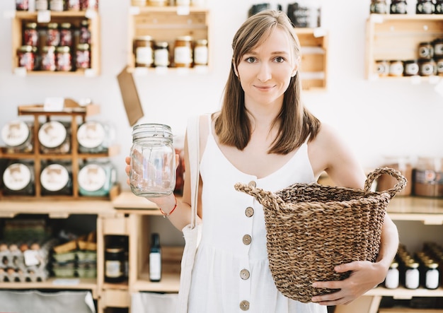 Mujer con vestido de lino blanco con frasco de vidrio vacío comprando en una tienda de comestibles libre de plástico sostenible Chica con una bolsa de algodón reutilizable y una cesta de mimbre en una tienda de desperdicio cero Estilo de vida minimalista de bajo desperdicio