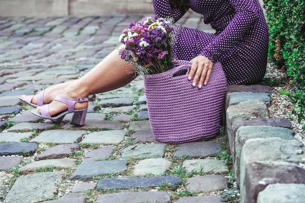 Mujer con vestido de lunares morados sosteniendo una bolsa de punto con flores posando en la calle de la ciudad