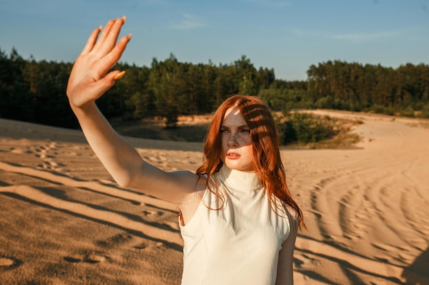 Foto mujer con vestido de moda que cubre la cara de la luz del sol mientras está de pie en las dunas de arena