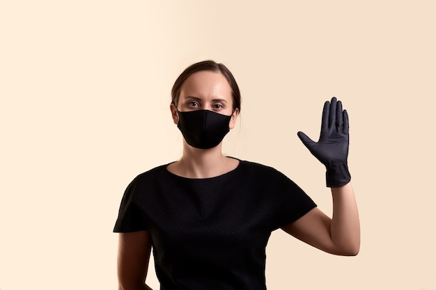 Foto mujer en vestido negro mascarilla y guantes muestra una palma como saludo y sobre pared beige