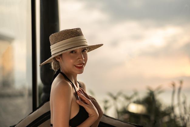 Mujer con vestido negro y sombrero de paja de pie en el balcón del hotel con vistas al atardecer