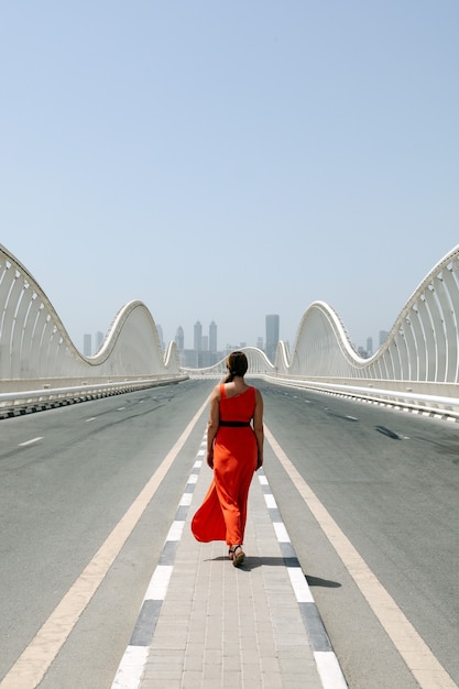 Una mujer en vestido rojo caminando por una carretera vacía en un puente Meydan con vistas a la ciudad al fondo