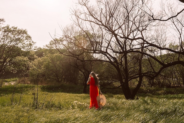 Una mujer con un vestido rojo se encuentra en un campo con un árbol al fondo.