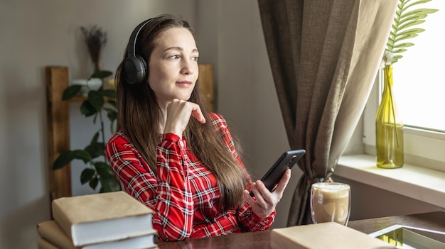 Una mujer con un vestido rojo está escuchando un audiolibro con auriculares y bebiendo café junto a la ventana. Concepto de educación tecnológica moderna y estudio de libros con alegría.