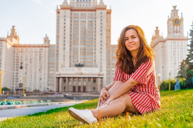 Una mujer con un vestido rojo se sienta en la hierba verde en frente del edificio principal de la Universidad de Moscú