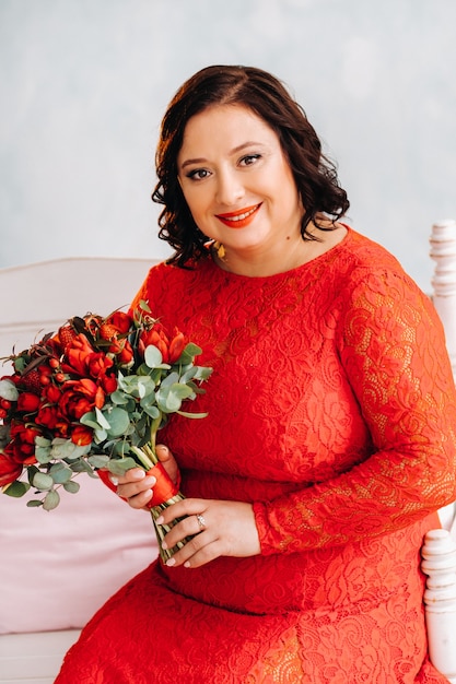 Una mujer con un vestido rojo se sienta en un sofá y sostiene un ramo de rosas rojas y bayas en el interior.