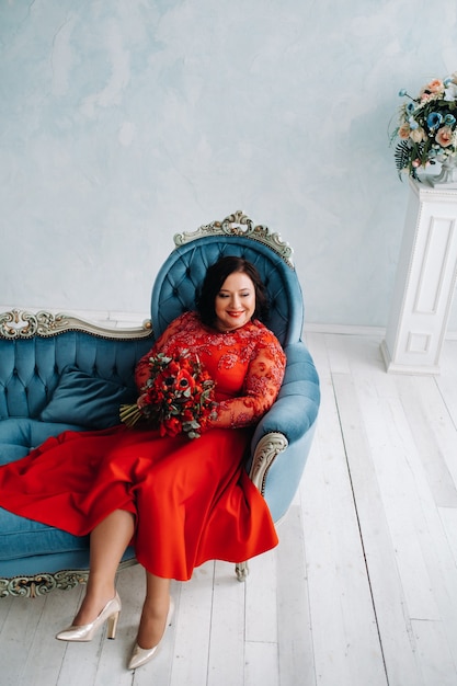 Una mujer con un vestido rojo se sienta en un sofá y sostiene un ramo de rosas rojas y fresas en el interior.