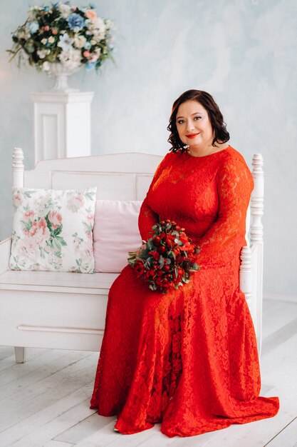 Una mujer con un vestido rojo se sienta en un sofá y sostiene un ramo de rosas rojas y fresas en el interior.