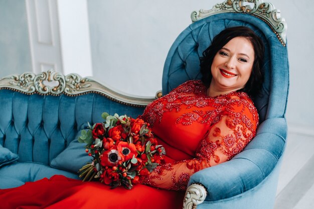 Una mujer con un vestido rojo se sienta en un sofá y sostiene un ramo de rosas rojas y fresas en el interior.