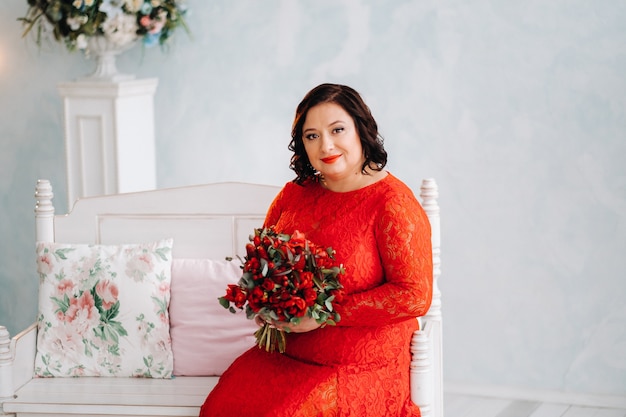 Una mujer con un vestido rojo se sienta en un sofá y sostiene un ramo de rosas rojas y fresas en el interior.