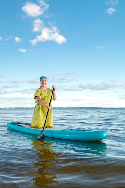 Una mujer con un vestido y un tocado de pie sobre una tabla de SUP con un remo flota en el agua contra el cielo azul