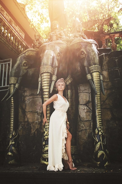 Mujer con vestido de túnica blanca y pañuelo en la cabeza posando junto a la estatua del elefante de tres cabezas