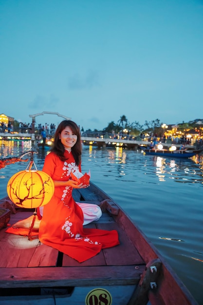 Mujer con vestido vietnamita Ao Dai, viajera en barco turístico y linterna de papel flotante en Hoi An, un antiguo hito de la ciudad para las atracciones turísticasVietnam y el concepto de viaje del sudeste