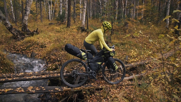 La mujer viaja en cicloturismo de terreno mixto con bikepacking El viaje del viajero con bolsas de bicicleta Ropa deportiva en colores verdes negros El viaje en el mágico bosque de otoño Río puente de madera