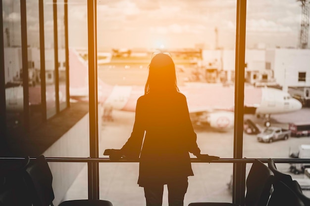 Mujer de viaje de pie viendo la puesta de sol en la ventana del aeropuerto.