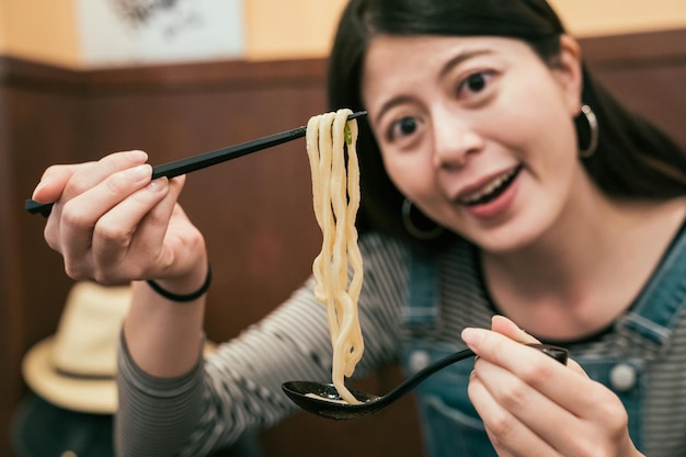 mujer viajera comiendo udon en un restaurante japonés para cenar. señora turista sosteniendo palillos y cuchara con comida sabrosa. niña sonriente mirando la cámara y mostrando deliciosos fideos en el interior.