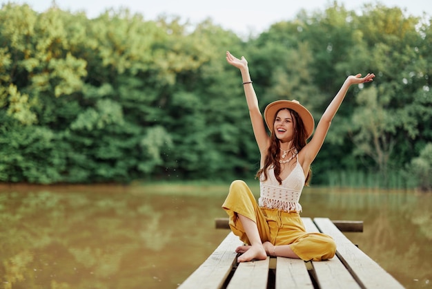 La mujer viajera ecoactivista hippie se sienta en un puente junto a un lago con los brazos extendidos con un sombrero y sonriendo sinceramente