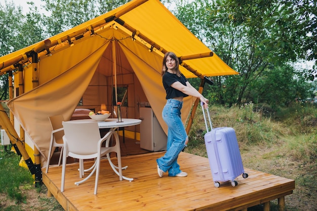 Mujer viajera feliz con equipaje de pie en el fondo de una acogedora casa glamping Tienda de campaña de lujo para vacaciones de verano al aire libre y vacaciones Concepto de estilo de vida