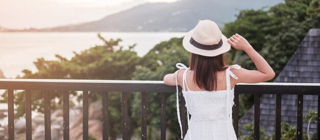 Mujer viajera feliz con vestido blanco y sombrero disfruta de hermosas vistas al mar mujer joven de pie en la playa tropical Libertad vacaciones relajantes vacaciones y concepto de viaje de verano