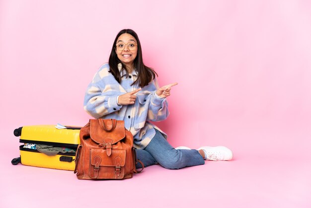 Mujer viajera con una maleta sentada en el suelo asustada y apuntando hacia un lado