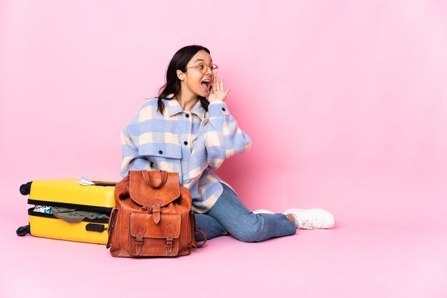 Mujer viajera con una maleta sentada en el suelo gritando con la boca abierta hacia un lado