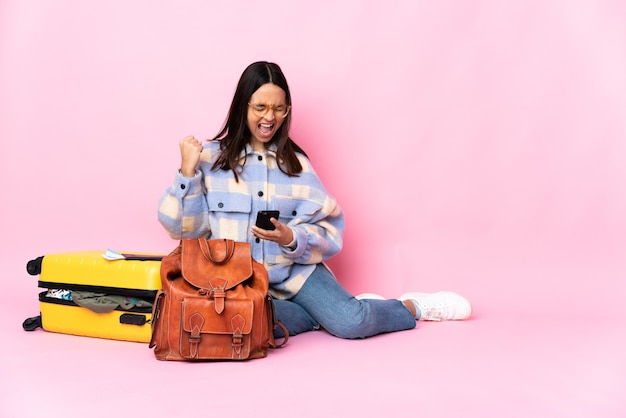 Mujer viajera con una maleta sentada en el suelo con teléfono en posición de victoria