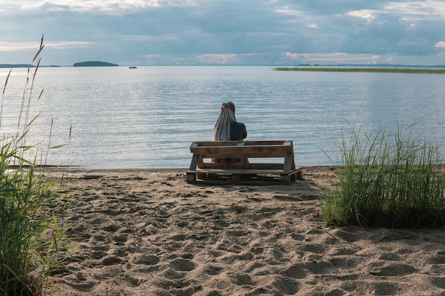 mujer viajera con peinado de trenzas sentada en un banco hecho de paletas viejas en una orilla de arena de un vasto lago