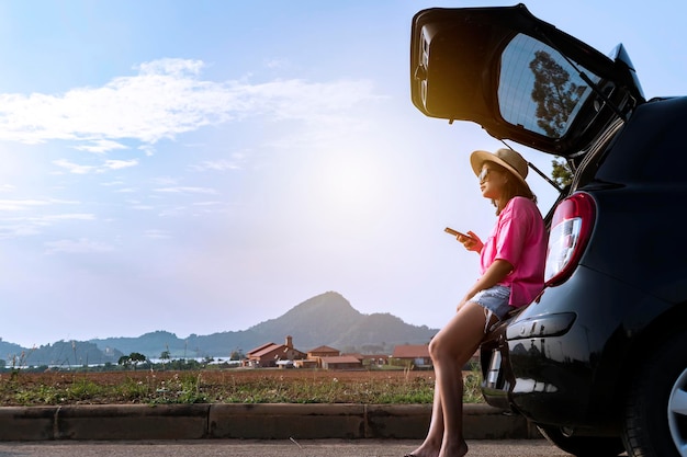 Mujer viajera sentada en un auto hatchback con fondo de montaña en tono vintage Ella está usando un teléfono inteligente