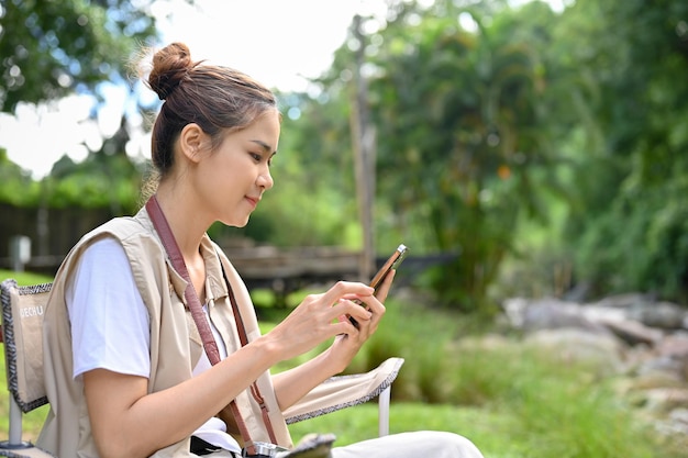 Mujer viajera sentada en una silla de camping y usando un teléfono inteligente para chatear con alguien