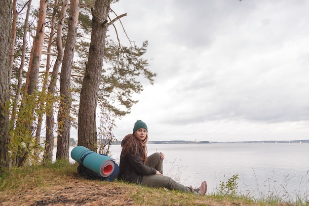 Mujer viajera solitaria sentada cerca del lago del bosque y mirando lejos.