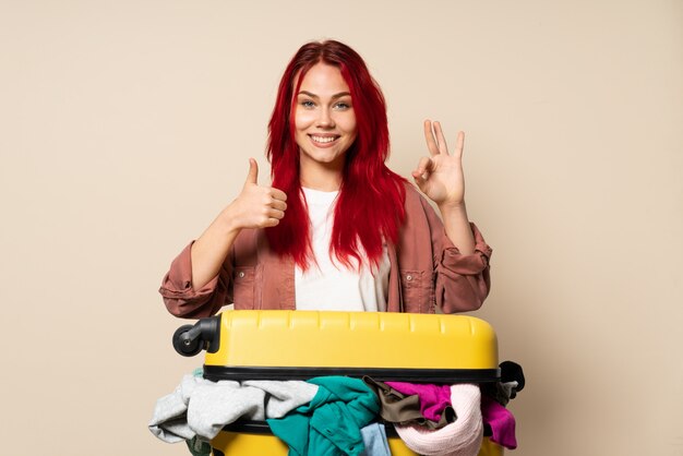 Mujer del viajero con una maleta llena de ropa aislado en la pared de color beige que muestra signo bien y gesto pulgar arriba